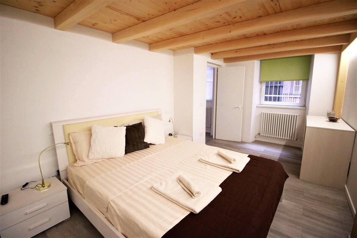 Quarto com cama, mesa de cabeceira e janela com persianas.