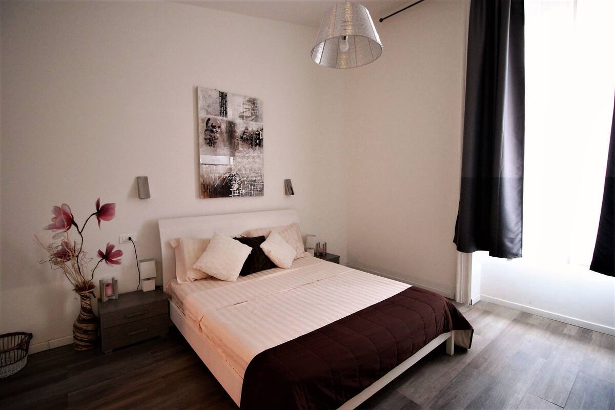 Quarto moderno com uma cama grande, uma mesa de cabeceira, uma luminária na parede e uma janela com cortinas.