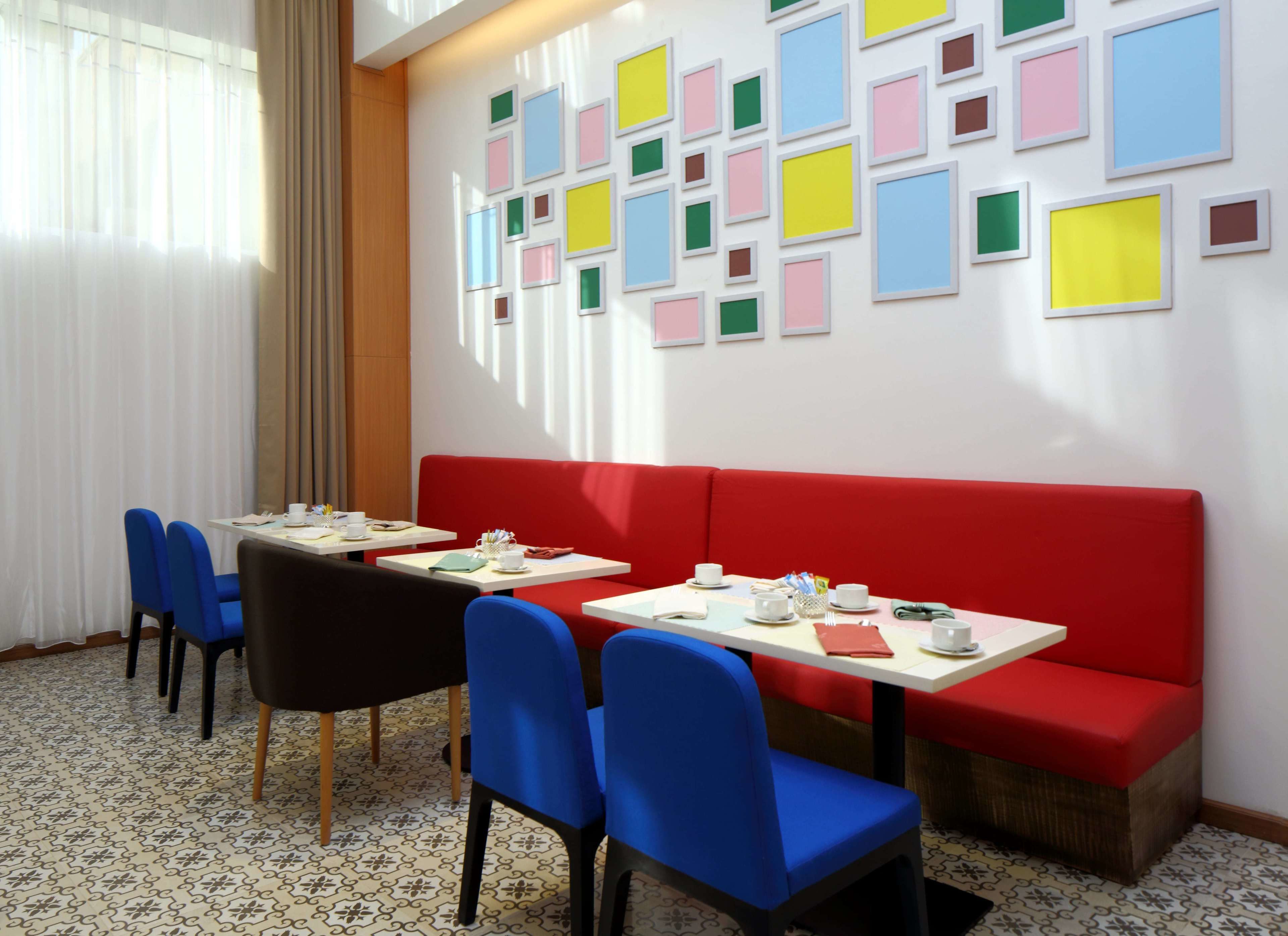 Un comedor con un banco rojo, sillas azules y mesas dispuestas con platos, tazas y cubiertos blancos.