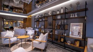 Um interior moderno com área de estar, sofás em tons claros e uma mesa redonda, cercada por estantes pretas com objetos decorativos e quadros.