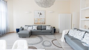 A modern living room with a grey sectional sofa, a white armchair, and a grey sofa with patterned cushions. There is a large abstract painting on the wall, a chandelier, and a white wardrobe. - Budapest Easy Flat - Basilica Lux (Budapest)