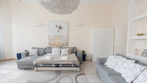 A modern bedroom with a bed, a grey sofa, a large abstract painting, and a chandelier. - Budapest Easy Flat - Basilica Lux (Budapest)