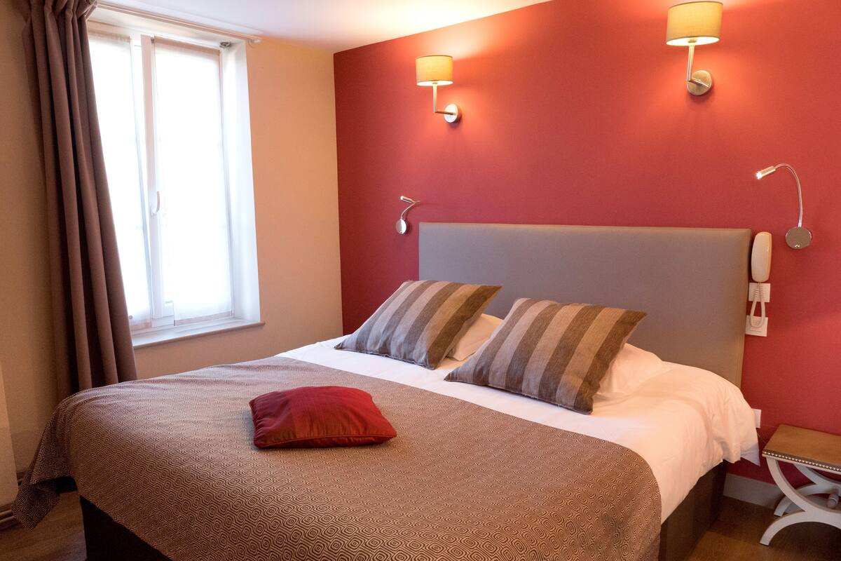 Ein Hotelzimmer mit einem großen Bett, zwei Kissen, einem roten Zierkissen, einem Nachttisch mit Lampe und einem Telefon an der Wand.