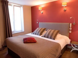 Ein Hotelzimmer mit einem großen Bett, zwei Kissen, einem roten Zierkissen, einem Nachttisch mit Lampe und einem Telefon an der Wand.