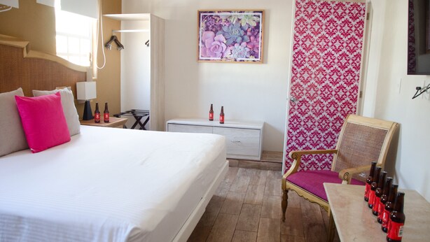 A hotel room with a bed, a chair, a table with bottles, a nightstand, a lamp, a painting, and a patterned curtain. - Santuario Playa (Playa del Carmen)