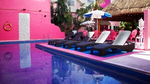 A swimming pool with lounge chairs and umbrellas, surrounded by pink buildings and palm trees. - Santuario Playa (Playa del Carmen)