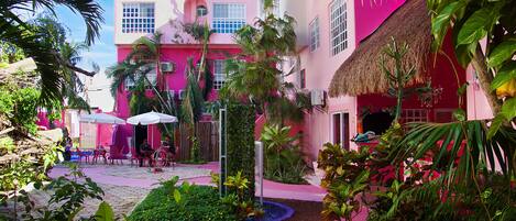 A vibrant courtyard with a pink building, outdoor seating, and lush greenery.