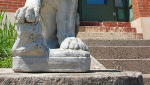 Uma estátua de leão de pedra em frente a um edifício de tijolos vermelhos, com porta e janelas verdes.