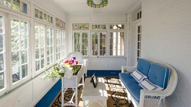 Uma sala iluminada pelo sol, com janelas amplas, um sofá de vime e uma mesa com um vaso de flores.