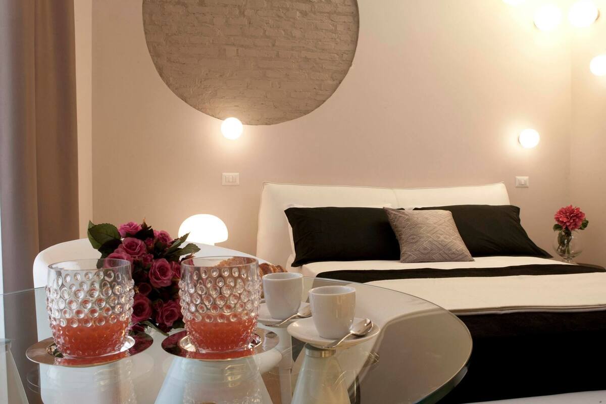 A hotel room with a bed, bedside table with glasses and a vase of flowers, and a round light fixture on the ceiling.