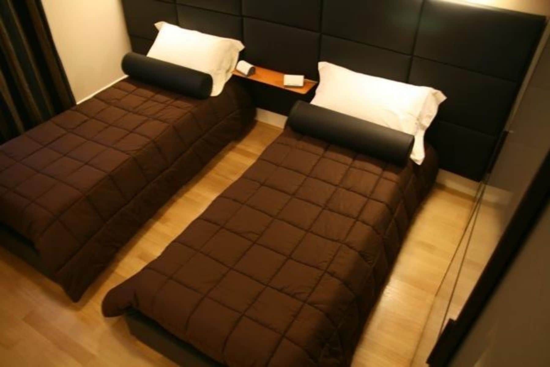 Deux lits simples avec une literie marron et des oreillers blancs, un plancher en bois et une tête de lit sombre.