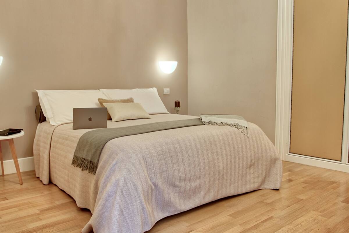 A bedroom with a bed, a laptop, a bedside table, and a wall-mounted light fixture.