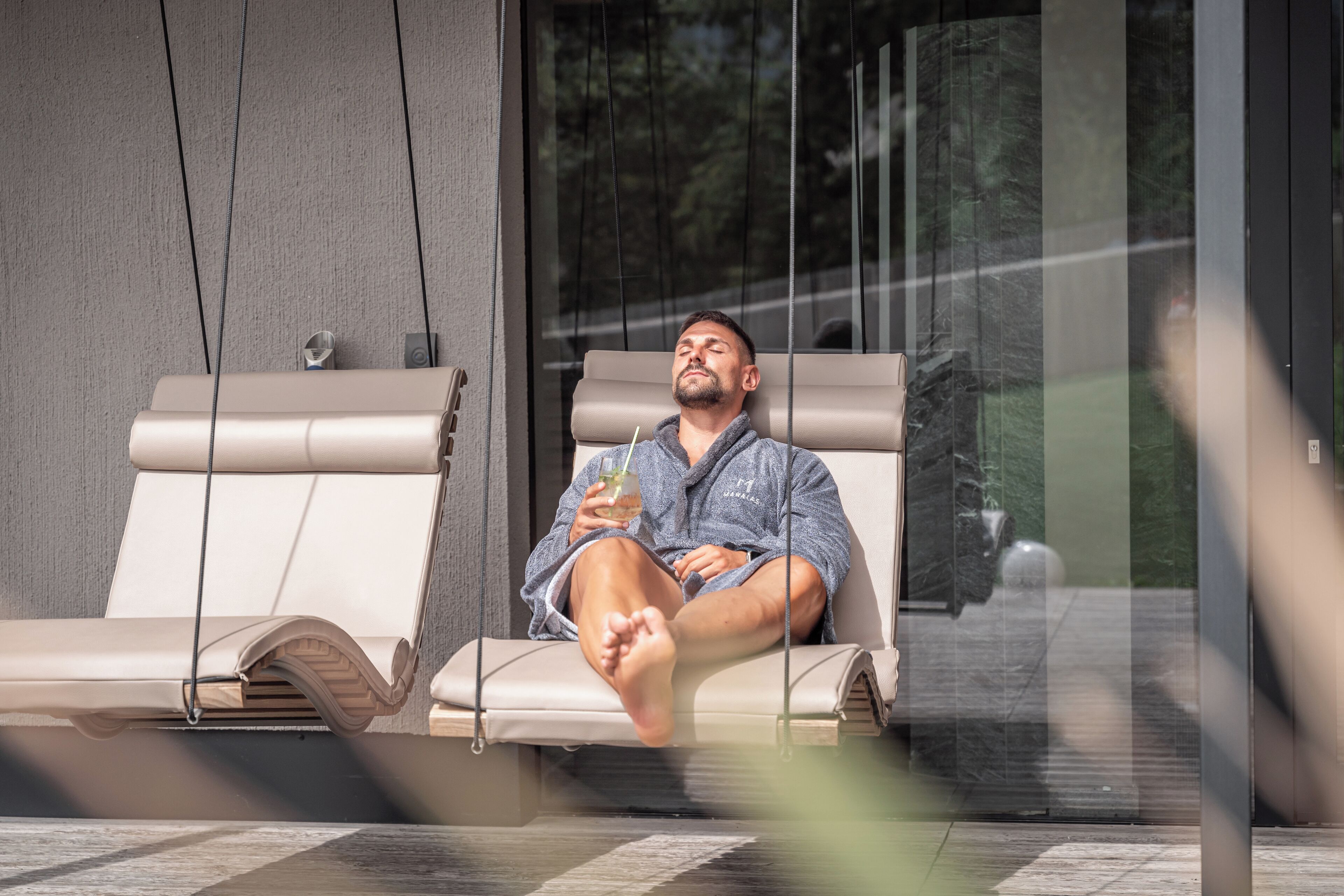 A person relaxing on a lounge chair with a drink, near a glass wall with a view of trees.