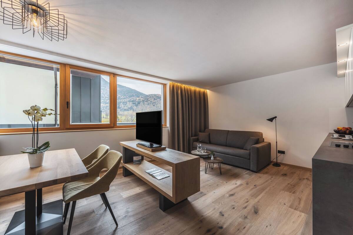 Un salon moderne avec un plancher en bois, un canapé, une table à manger avec des chaises, un téléviseur et une grande fenêtre avec des rideaux.