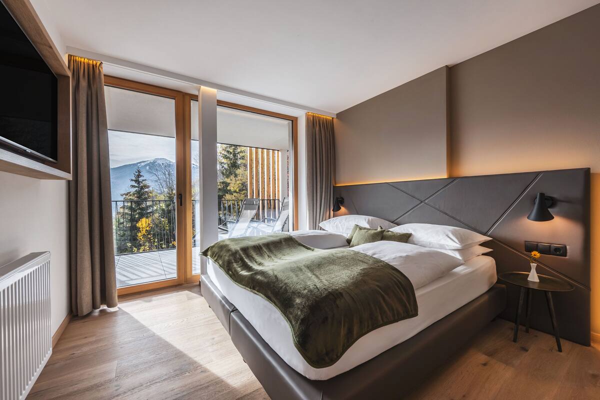 Une chambre d’hôtel moderne dotée d’un grand lit, d’un téléviseur fixé au mur, d’un balcon offrant une vue sur les montagnes et d’une petite table avec une lampe.