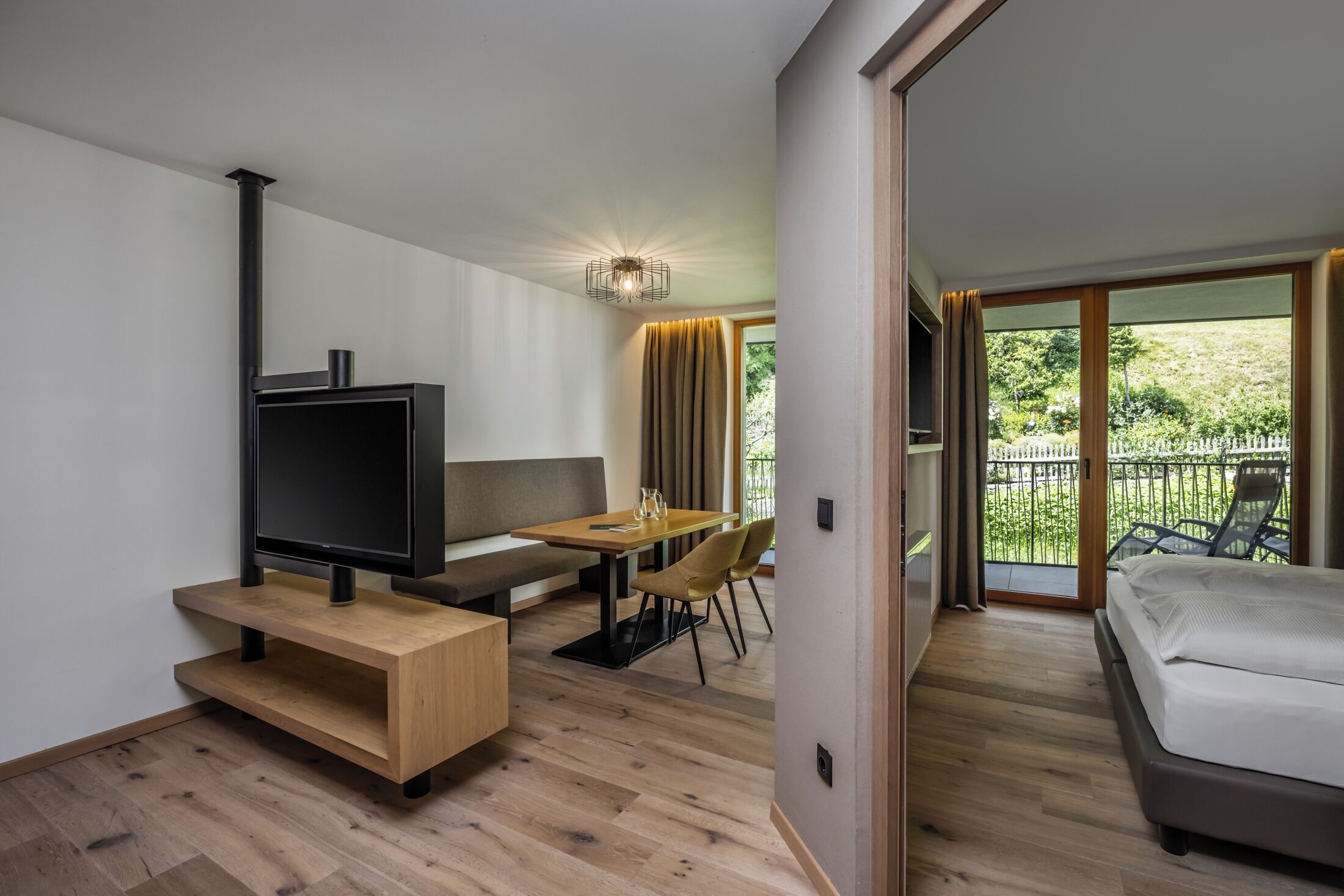 Une chambre d’hôtel moderne avec un plancher en bois, une télévision, un canapé, une table à manger avec des chaises et un balcon donnant sur un paysage verdoyant.