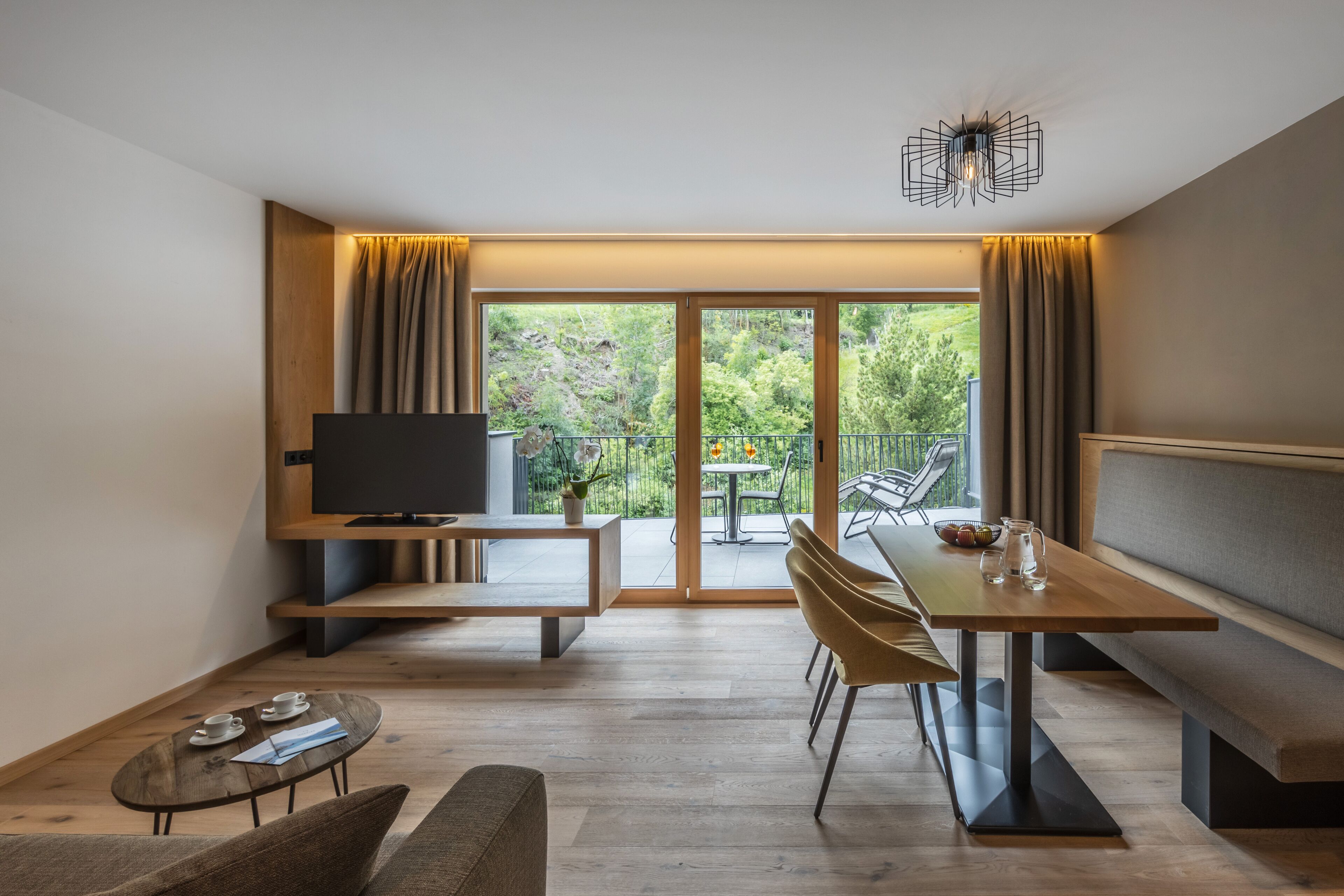 A modern living room with a TV, wooden furniture, a dining table, and a balcony with a view of greenery.
