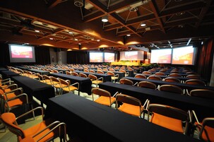 Un salón de conferencias con mesas y sillas dispuestas en filas, equipado con pantallas grandes para presentaciones.