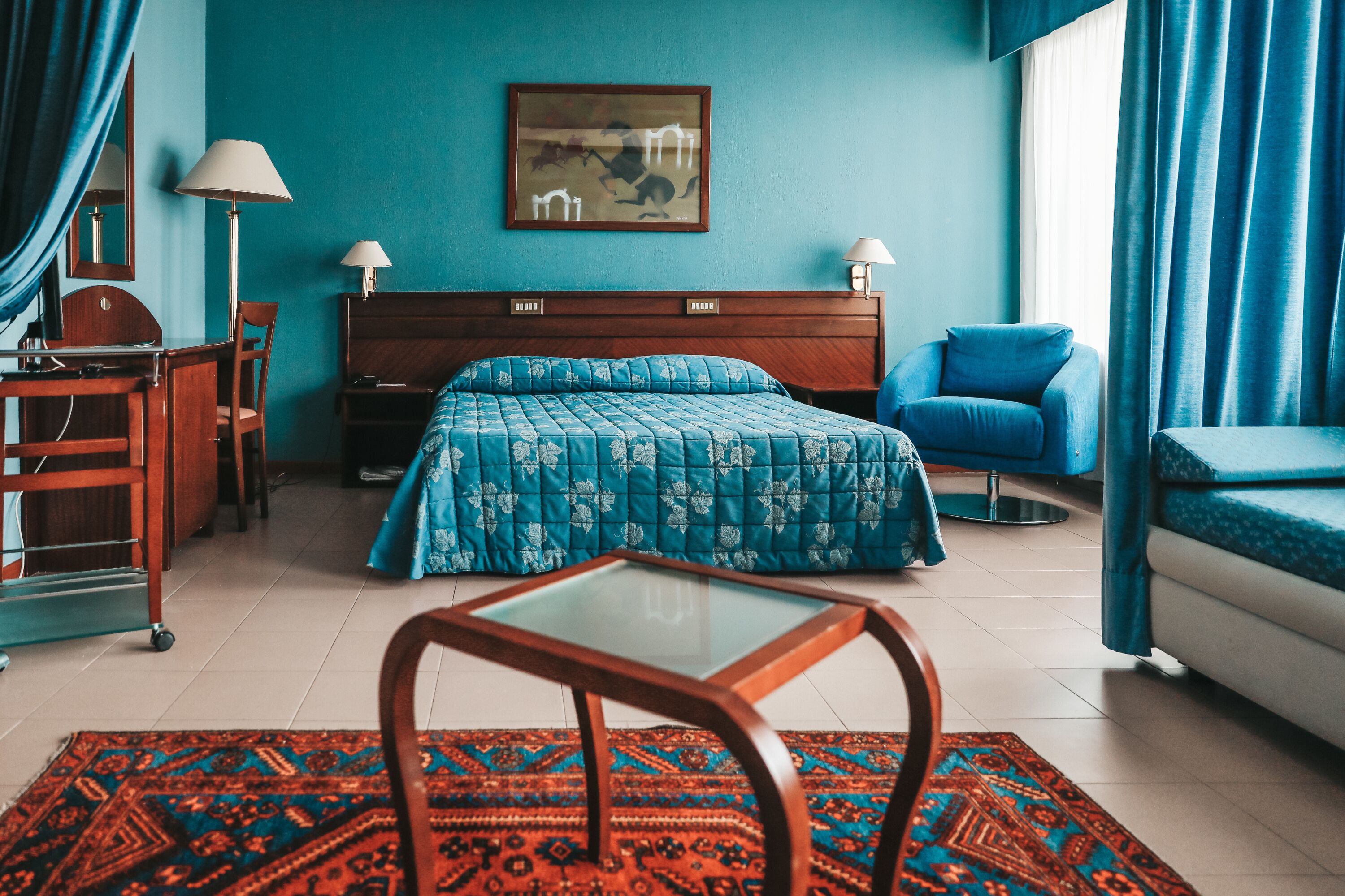 Una camera d'albergo con un letto, una scrivania, una sedia, un tavolino e una finestra con tende.