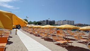 Una playa con filas de tumbonas y sombrillas amarillas, una bandera y edificios al fondo.