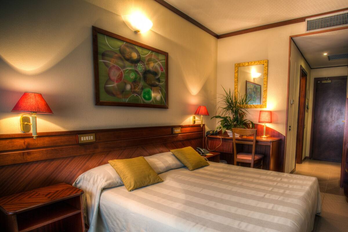 Une chambre d’hôtel avec un grand lit, deux tables de chevet, un bureau avec une chaise, une plante en pot et un tableau au mur.