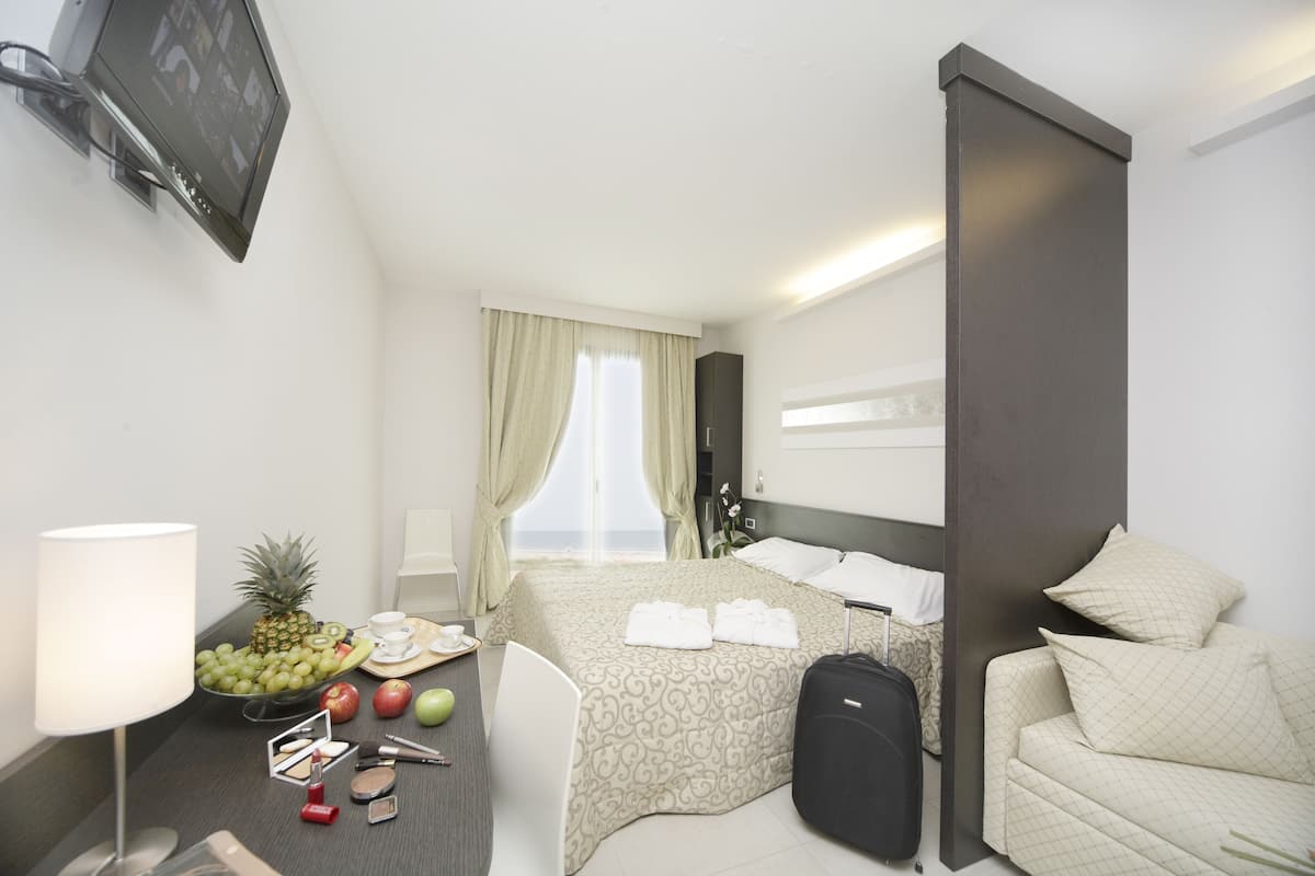 Une chambre d’hôtel avec un lit, un bureau avec une lampe, une chaise, une valise et un panier de fruits.