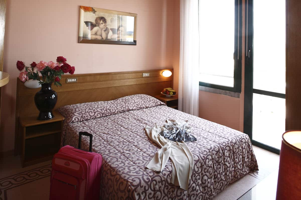 Une chambre d’hôtel avec un lit, une table de chevet avec une lampe, un vase avec des fleurs et une valise.