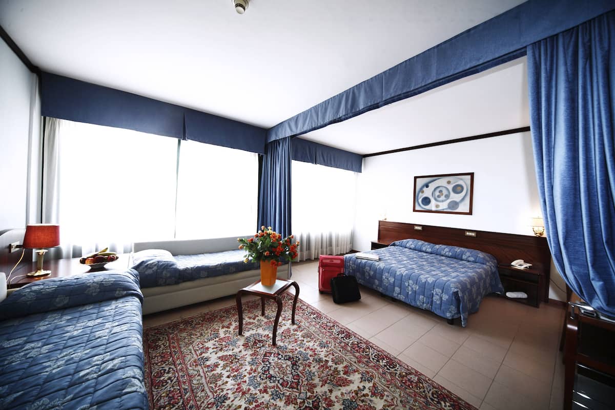 Une chambre d’hôtel avec deux lits, un coin salon, une grande fenêtre avec des rideaux, un tapis à motifs floraux et un tableau au mur.