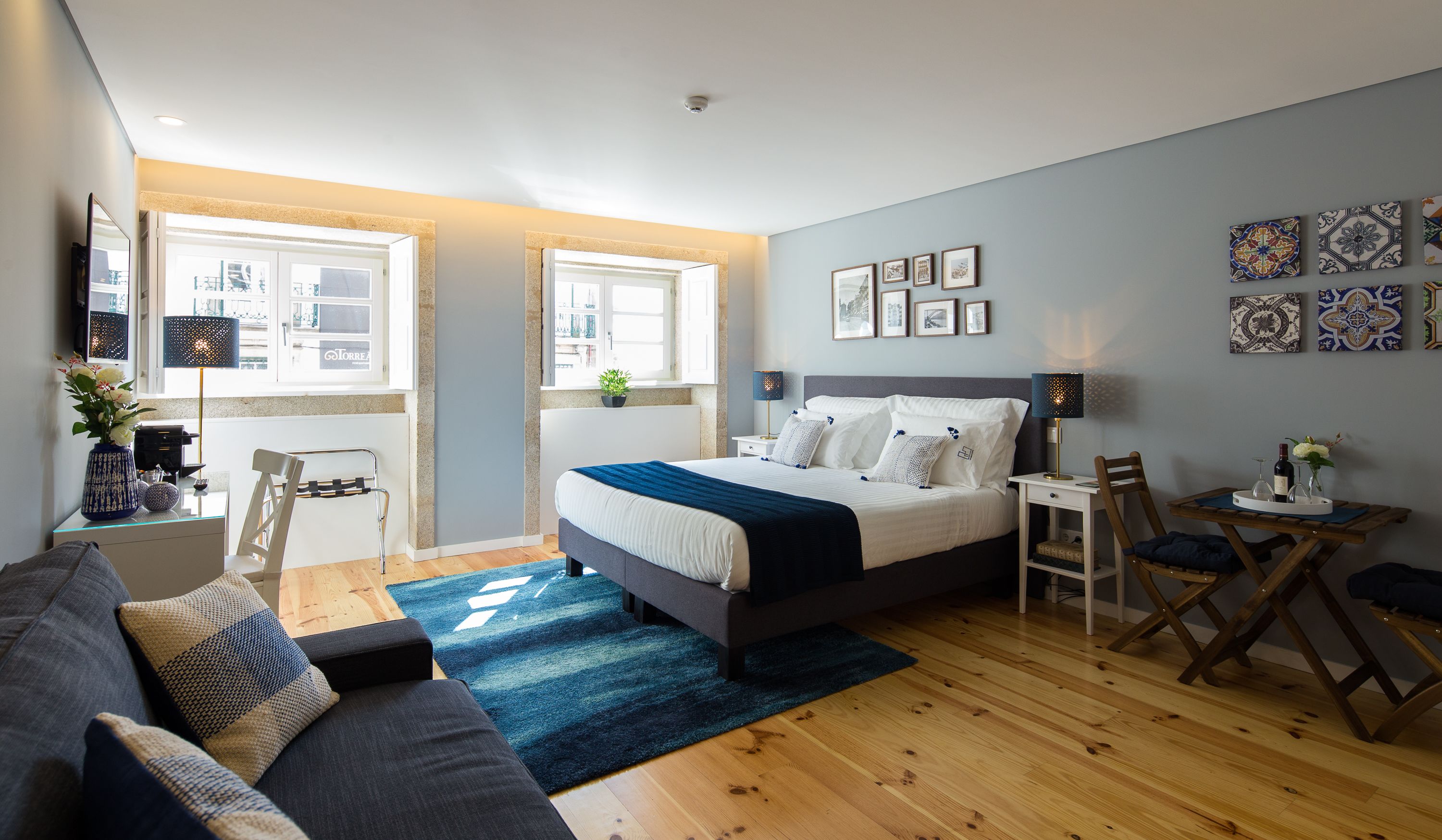Ein modernes Schlafzimmer mit einem großen Bett, einem Schreibtisch und einem Stuhl. Es gibt Fenster, eine Pflanze und Kunstwerke an den Wänden.