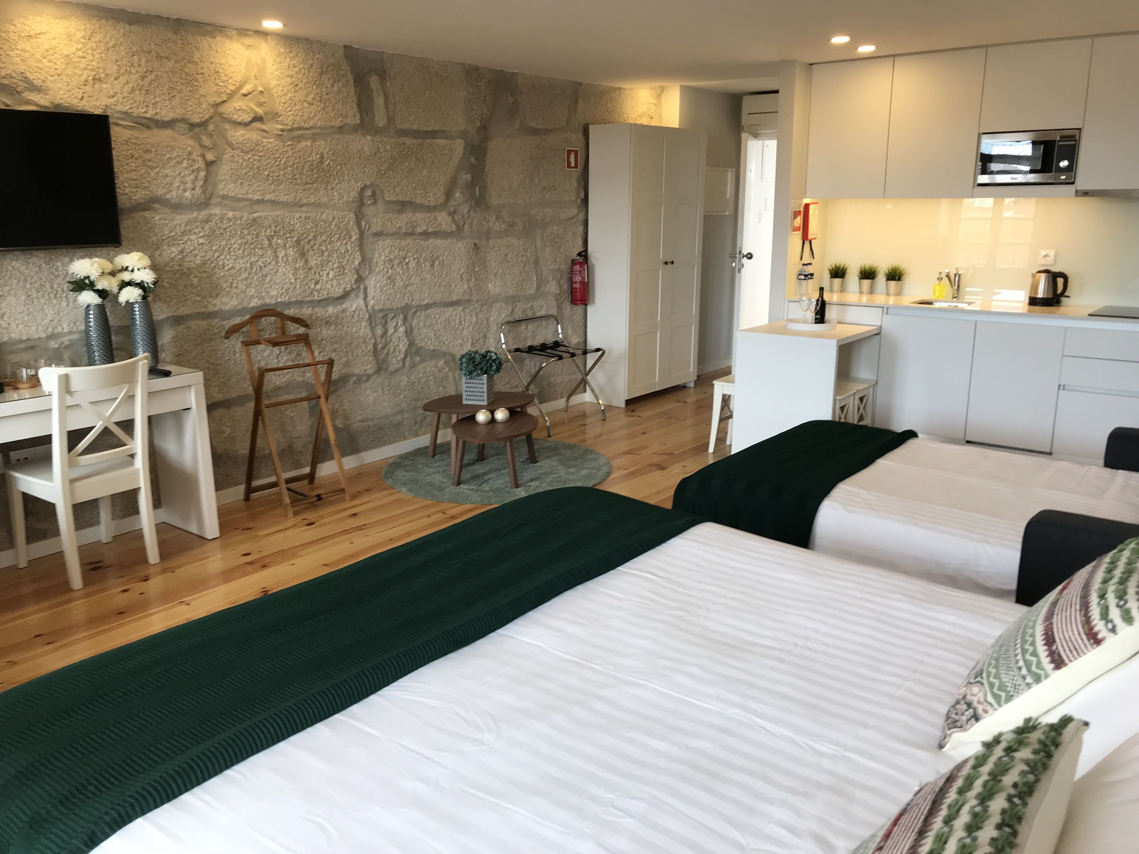 Una habitación moderna con pared de piedra, cama con ropa de cama verde y blanca, un escritorio con silla, una pequeña mesa con adornos y una kitchenette con microondas.