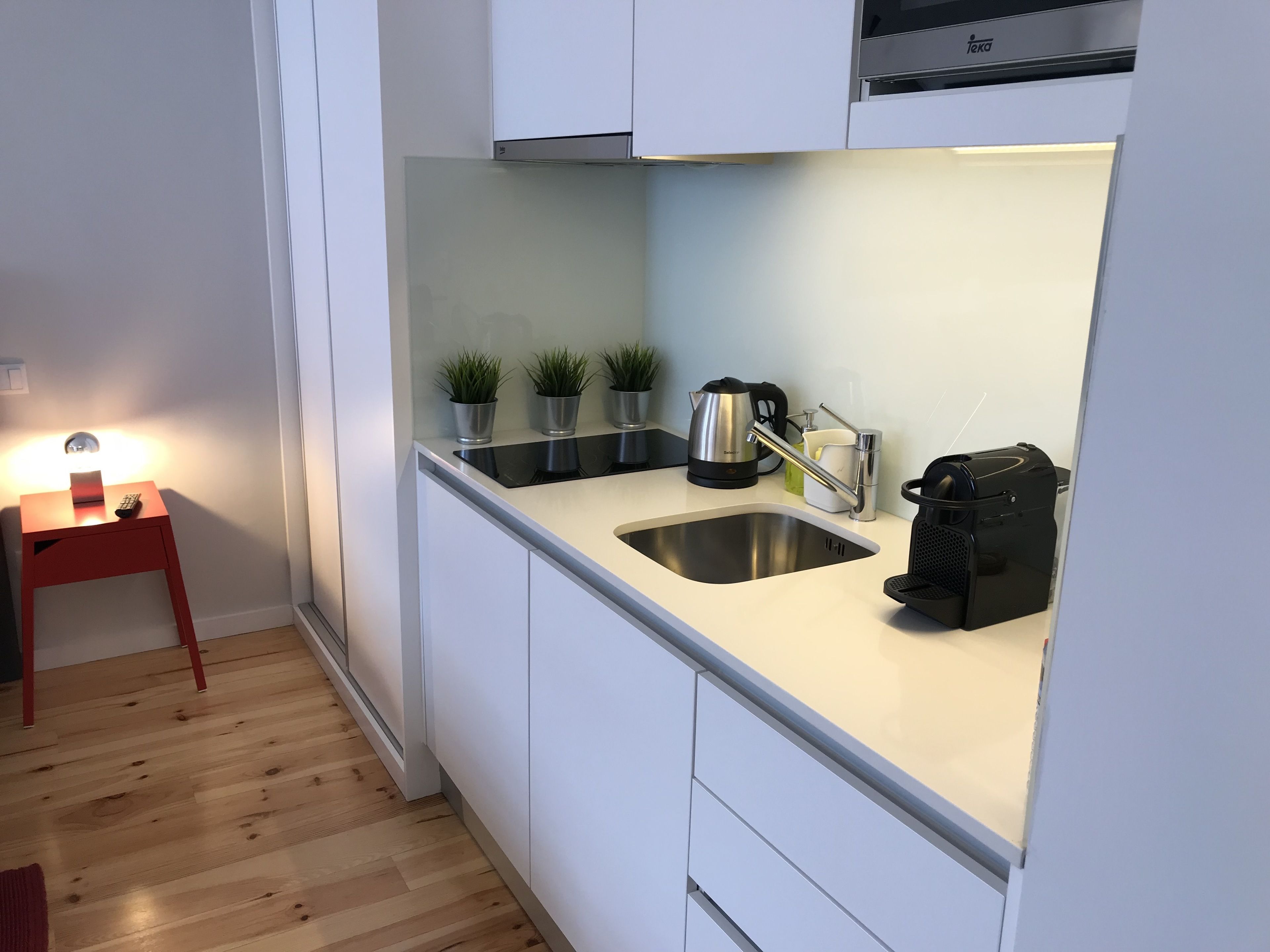 A modern kitchen with white cabinets, a stainless steel sink, a kettle, and a coffee machine. There are three potted plants on the countertop.