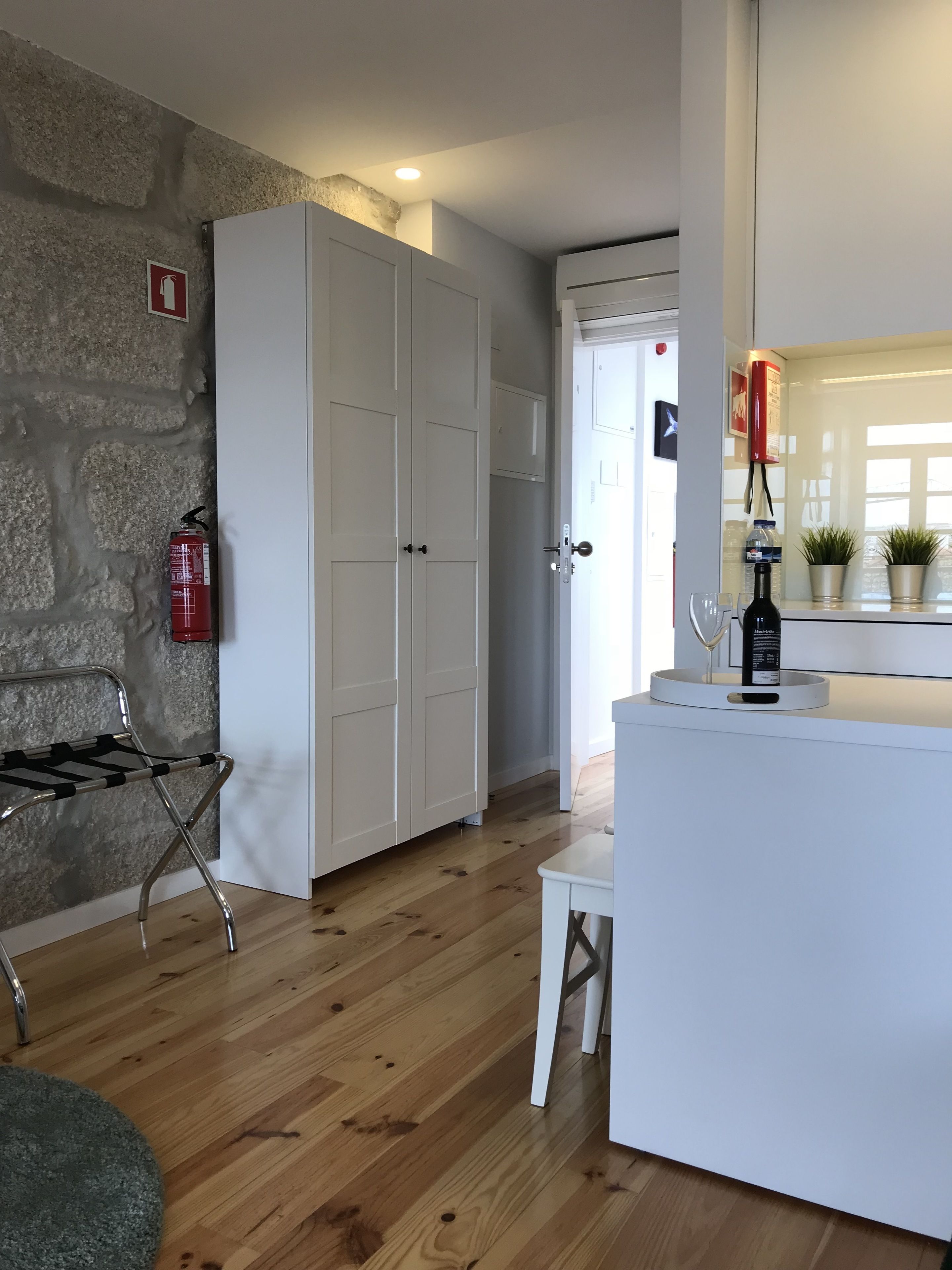 Een kamer met een stenen muur, een witte inbouwkast, een houten vloer, een brandblusser, een klein tafeltje met een fles en een deur die leidt naar een andere kamer.
