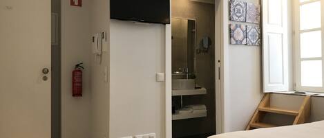 A hotel room with a bed, a TV mounted on the wall, a bathroom with a sink and mirror, and a fire extinguisher on the wall.