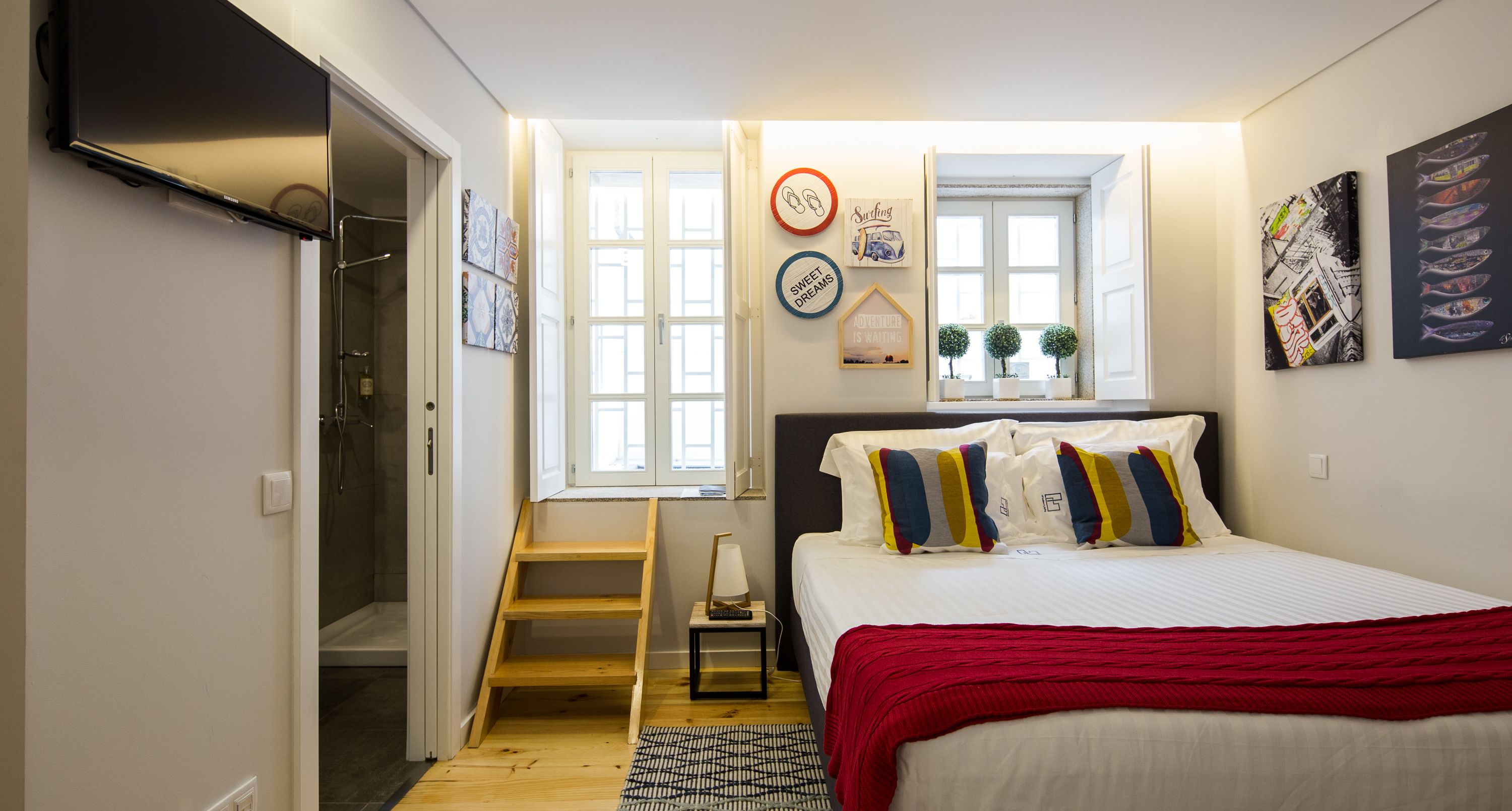 A bedroom with a bed, a red throw blanket, a wooden ladder, a small table with a lamp, and a window with a view of the outside.