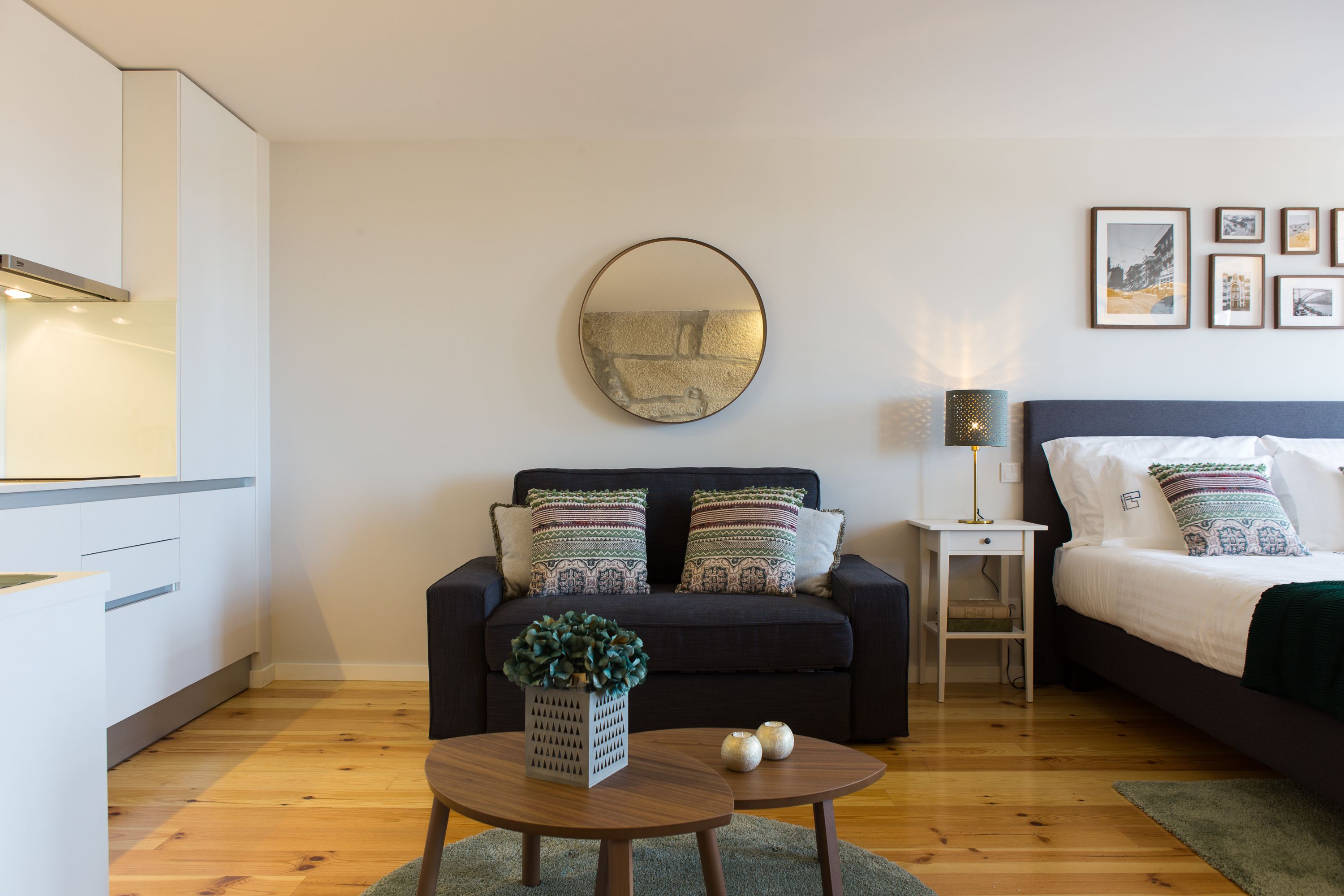 A modern bedroom with a dark sofa, a round mirror, a bedside table with a lamp, and a bed with patterned pillows.
