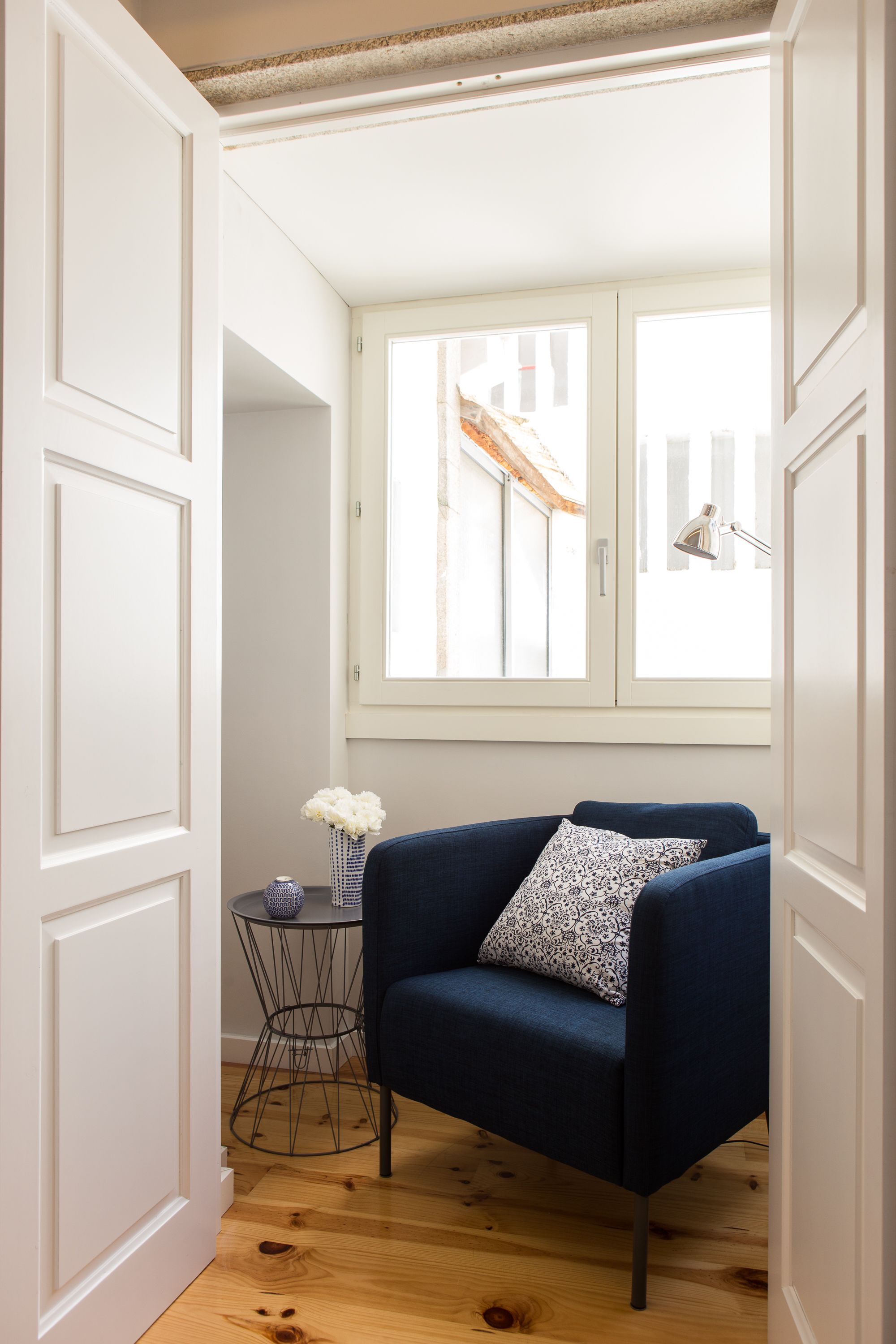 A room with a blue armchair, a small round table with a vase of flowers, and a window with a view of buildings.