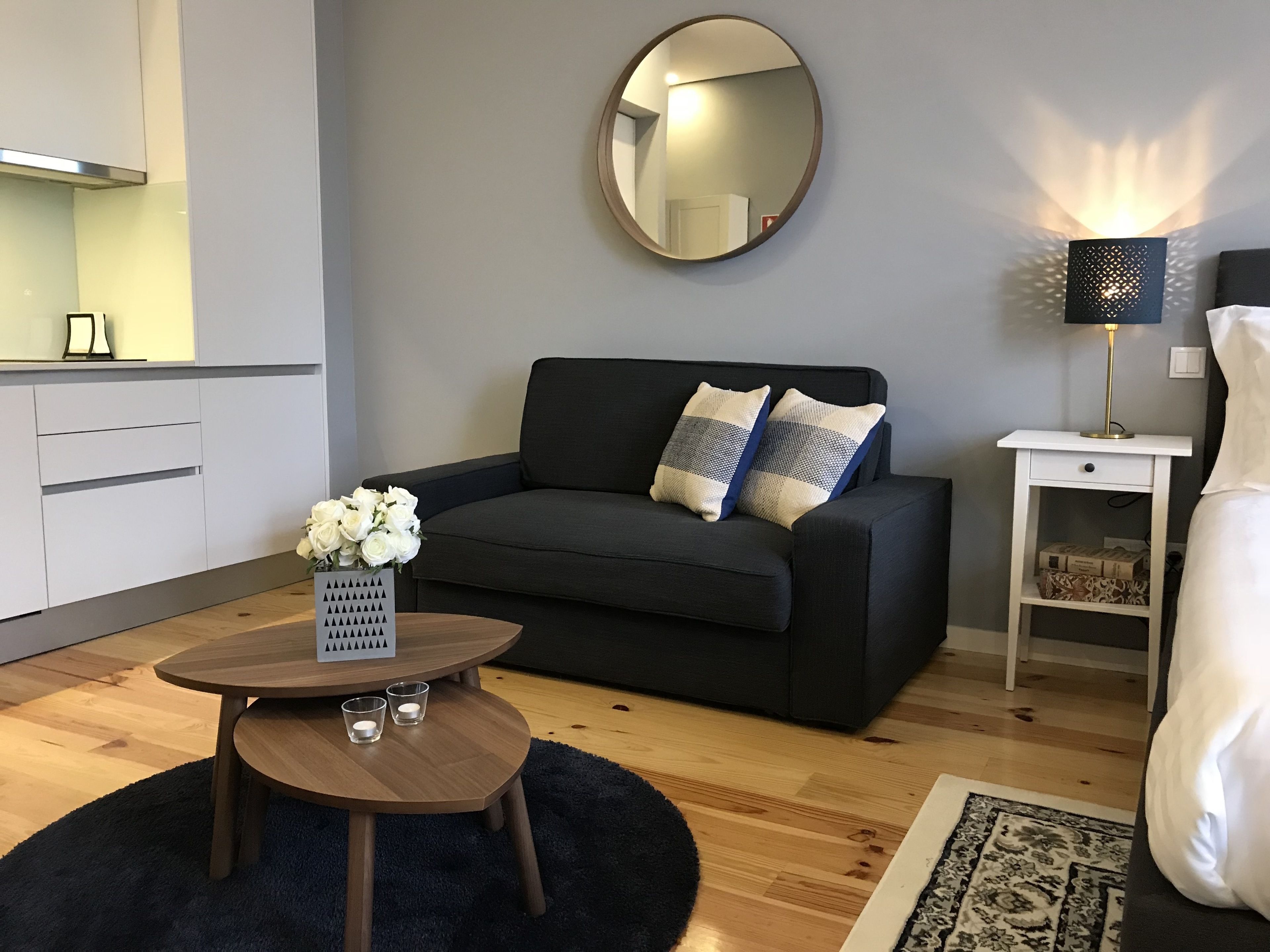 A modern living room with a dark grey sofa, a round mirror, a small round table with candles, a lamp, and a bed with white bedding.