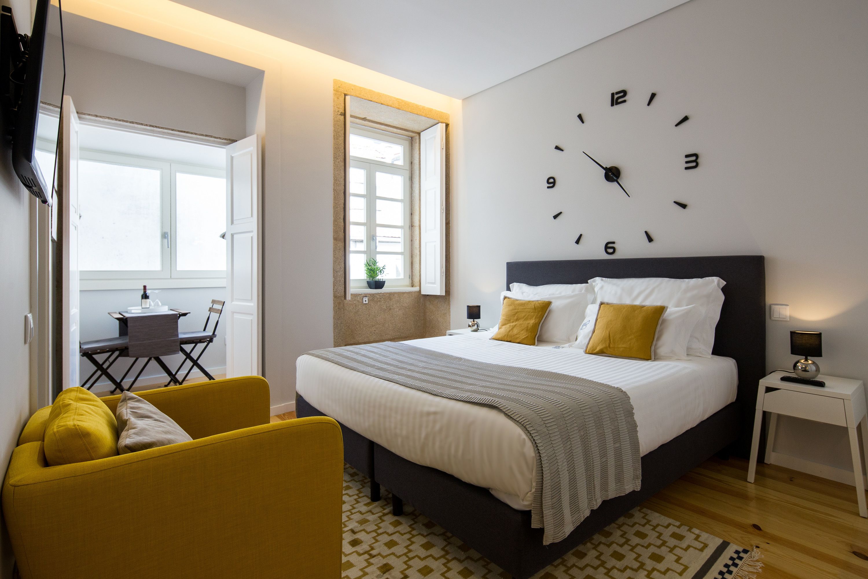 A modern hotel room with a large bed, a small desk, a yellow armchair, and a wall clock.