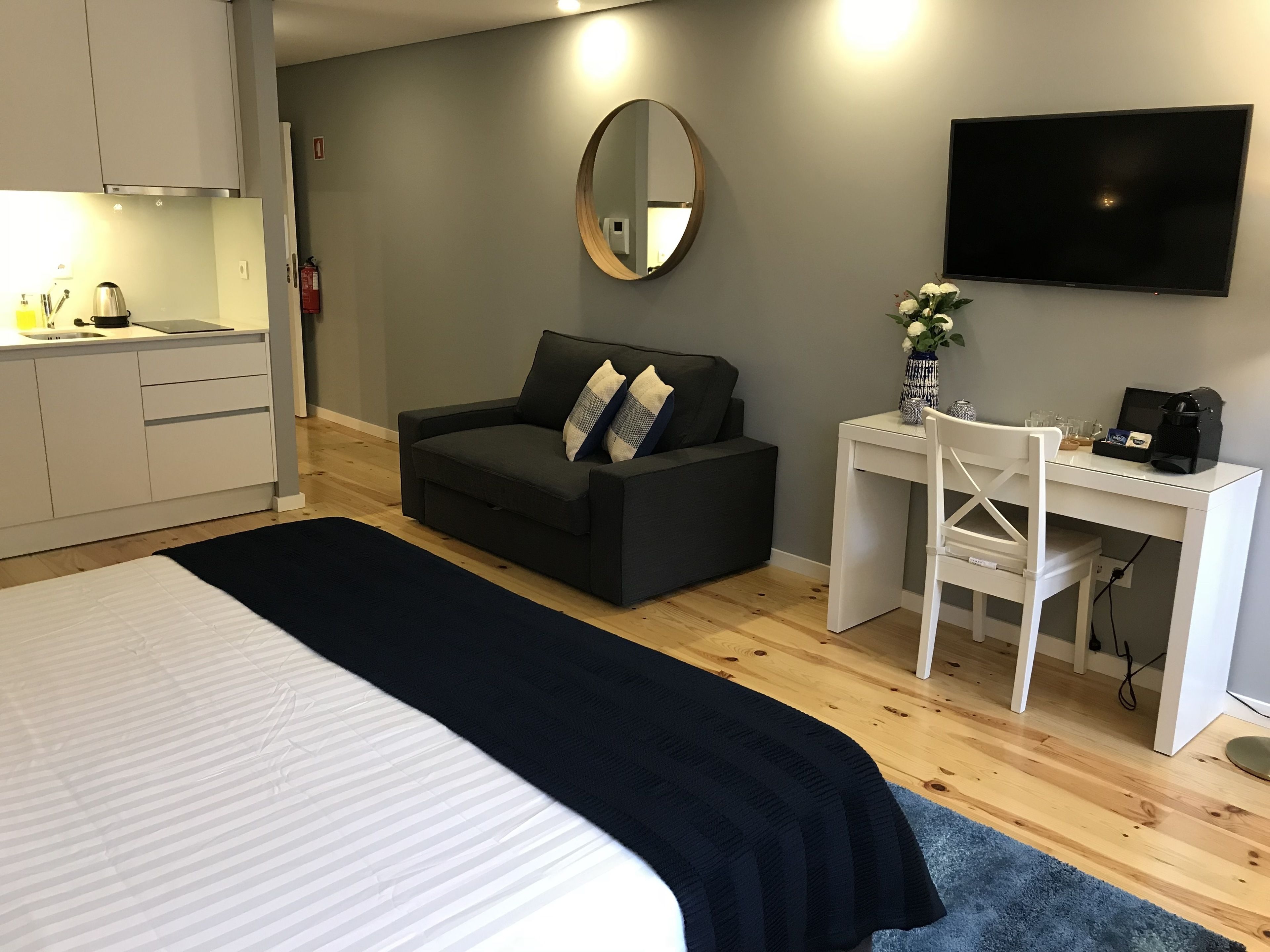 Ett modernt hotellrum med en stor säng, en mörkgrå soffa, ett vitt skrivbord med en stol, en platt-TV och ett litet bord med en vas med blommor.