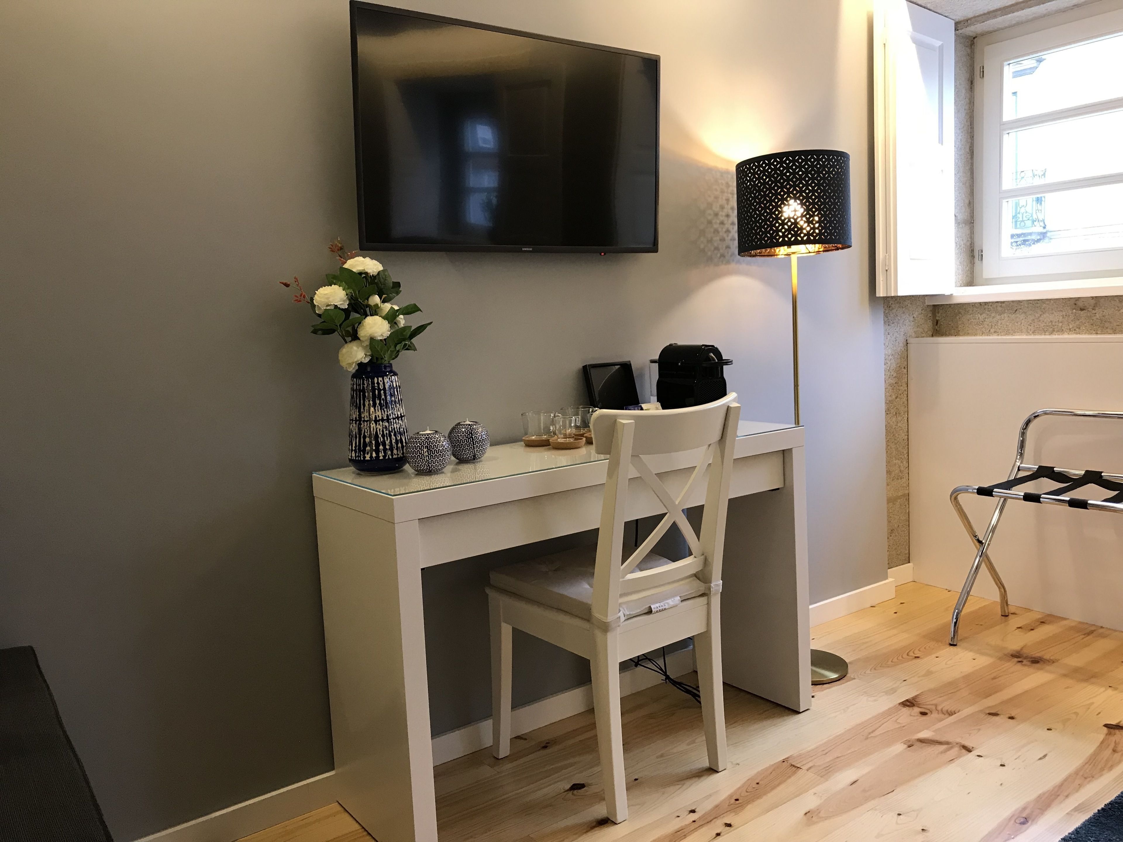 Una habitación pequeña y moderna con un escritorio blanco, una silla, un televisor montado en la pared, una lámpara y una ventana.
