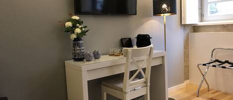 A small, modern room with a white desk, a chair, a TV mounted on the wall, a lamp, and a window.