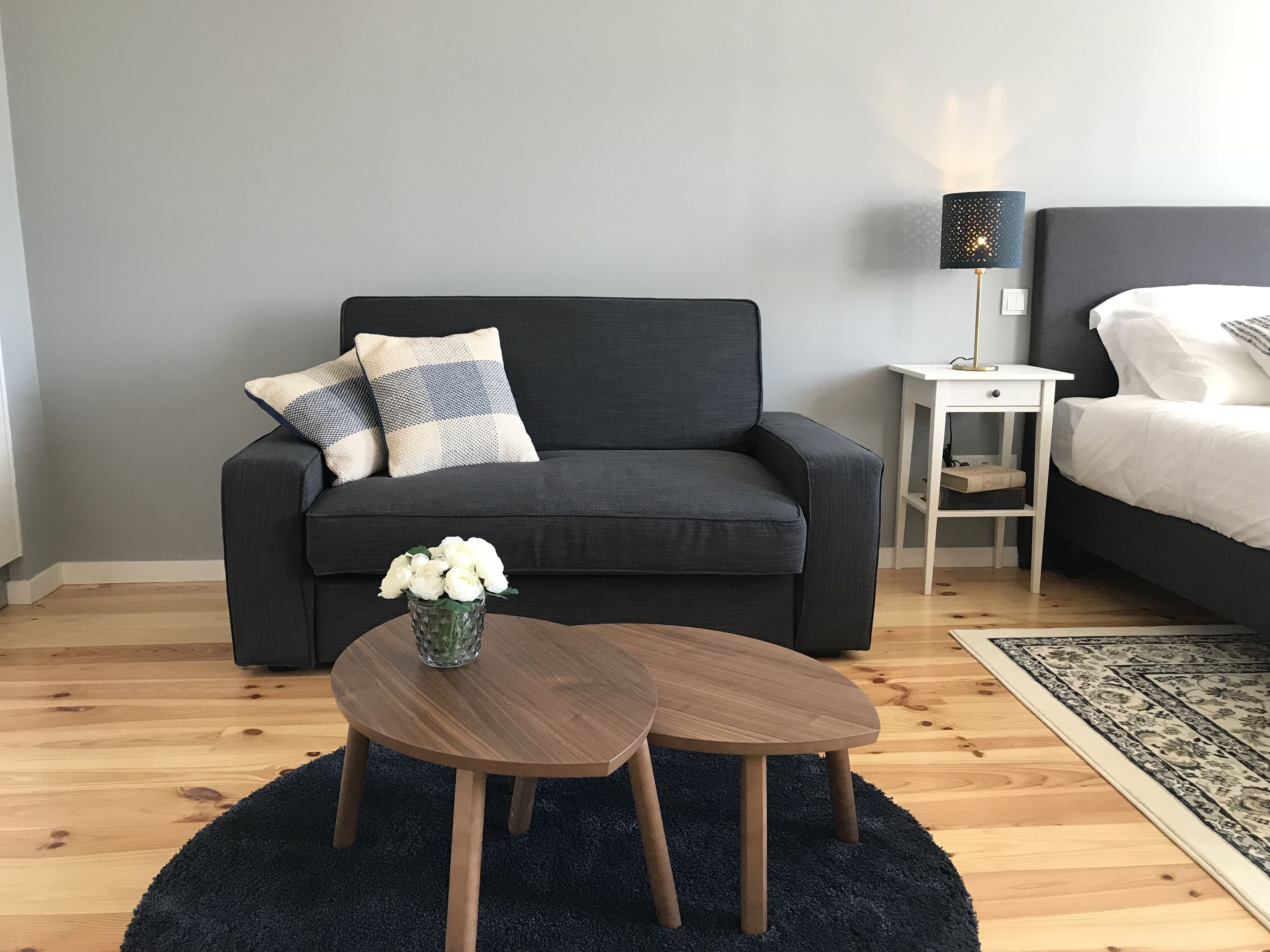 A modern bedroom with a grey sofa, a small bedside table, a lamp, and a bed with white bedding.