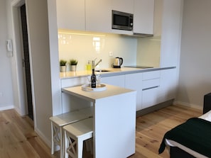 Cocina compacta con muebles blancos, microondas, lavamanos y un pequeño comedor con un banco.