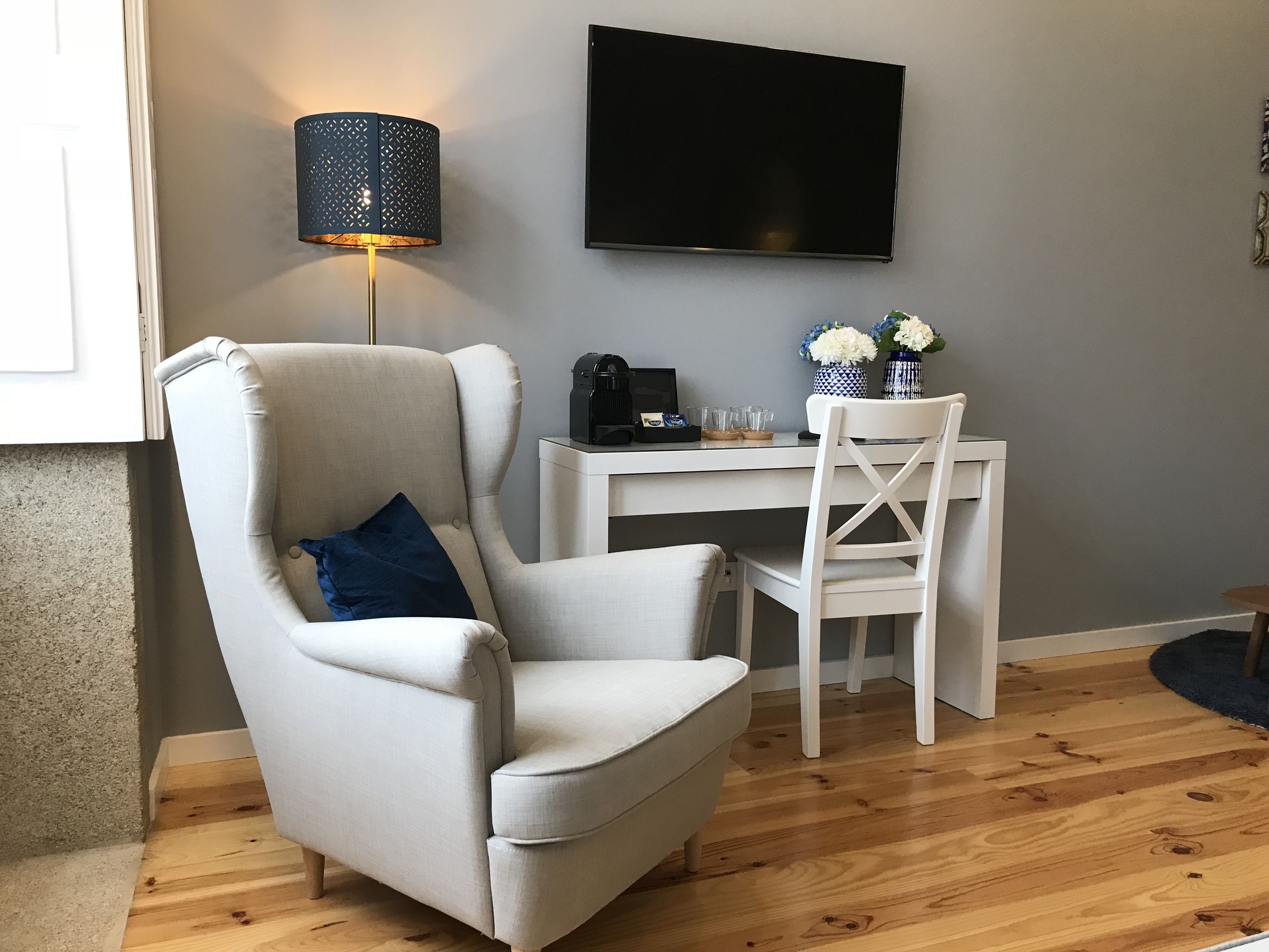 Een woonkamer met een beige fauteuil, een witte eettafel, een aan de muur bevestigde televisie en een lamp met een patroonlampenkap.