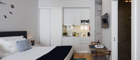 A modern bedroom with a bed, bedside table, and a small desk with a chair. There is a kitchen area with a microwave, sink, and cabinets. A TV is mounted on the wall above the kitchen.