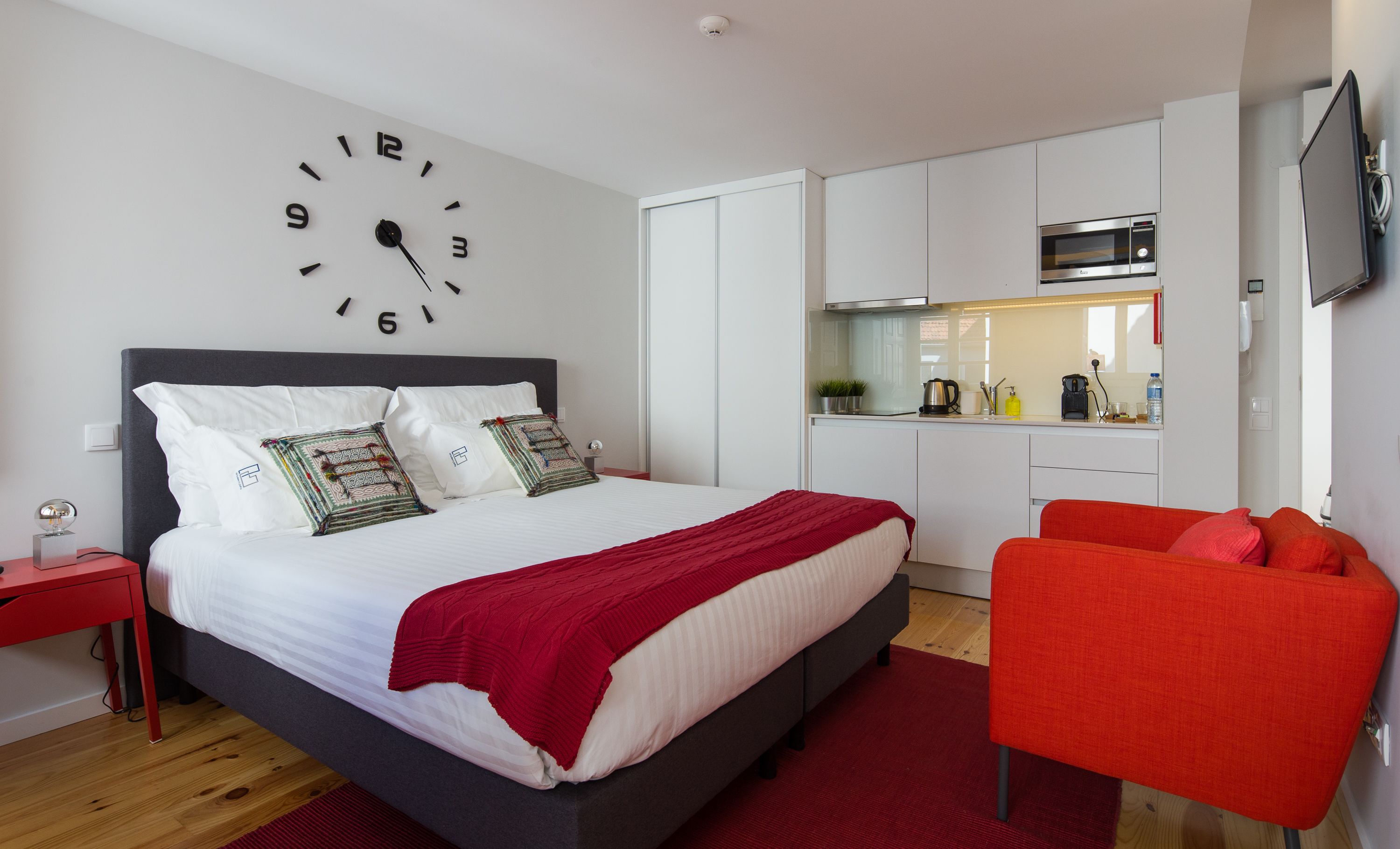 A modern bedroom with a bed, a red chair, a kitchen area, and a wall clock.
