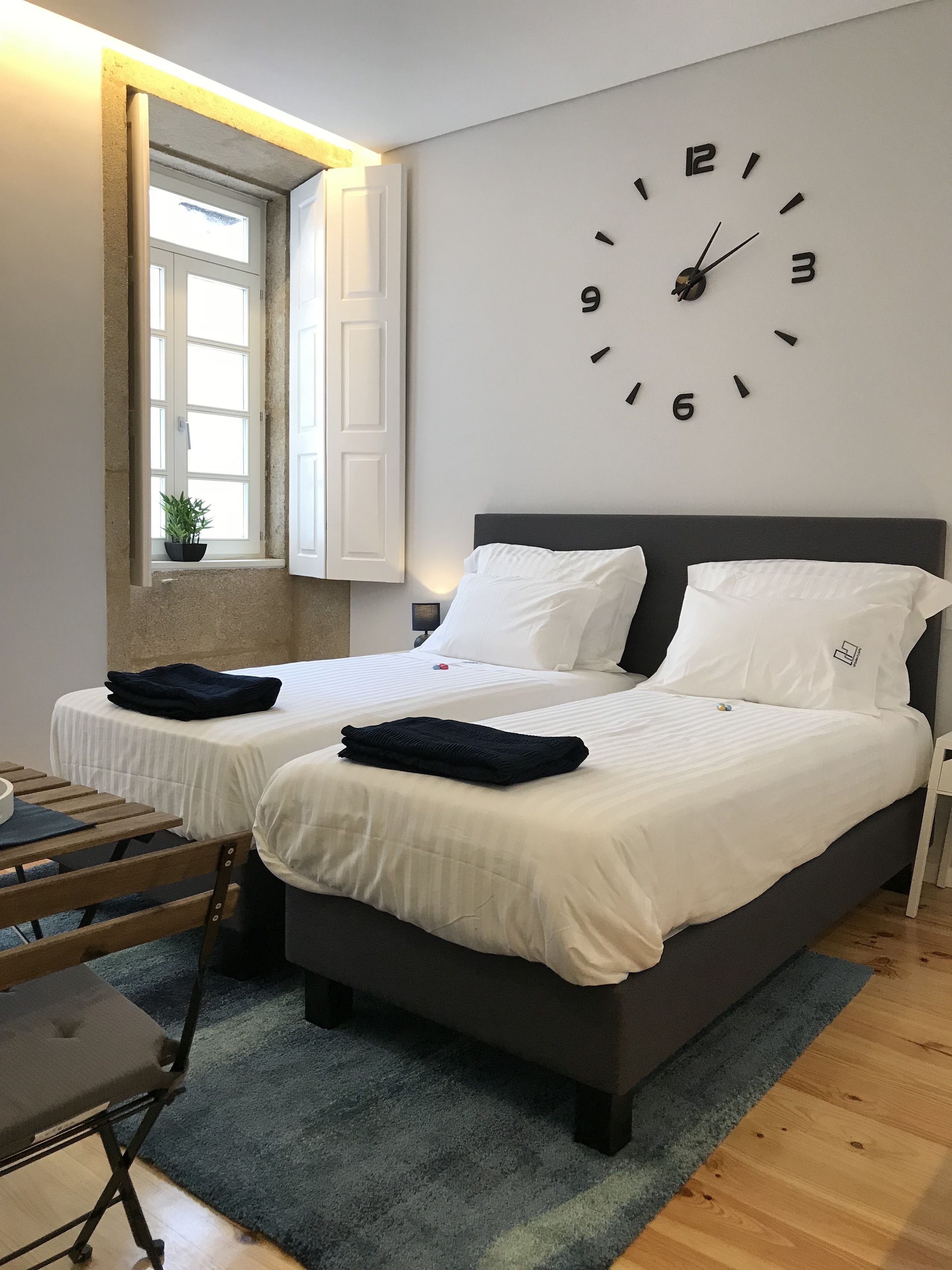 A bedroom with two beds, a large wall clock, a window with curtains, and a small table with a chair.