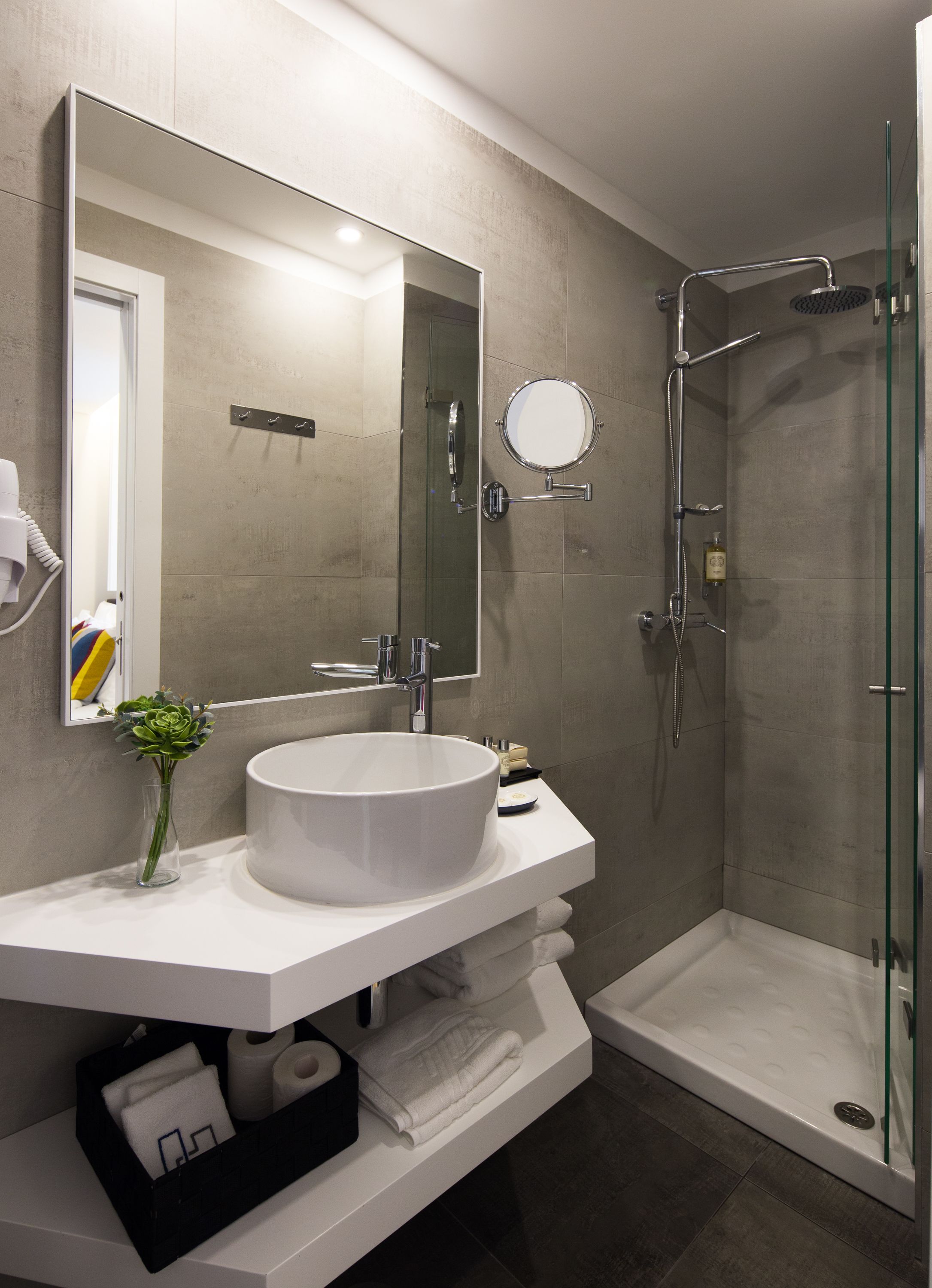 A modern bathroom with a large mirror, a round mirror, a white sink, a glass shower enclosure, and a small vase with green flowers.