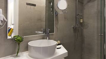 A modern bathroom with a large mirror, a round mirror, a white sink, a glass shower enclosure, and a small vase with green flowers.