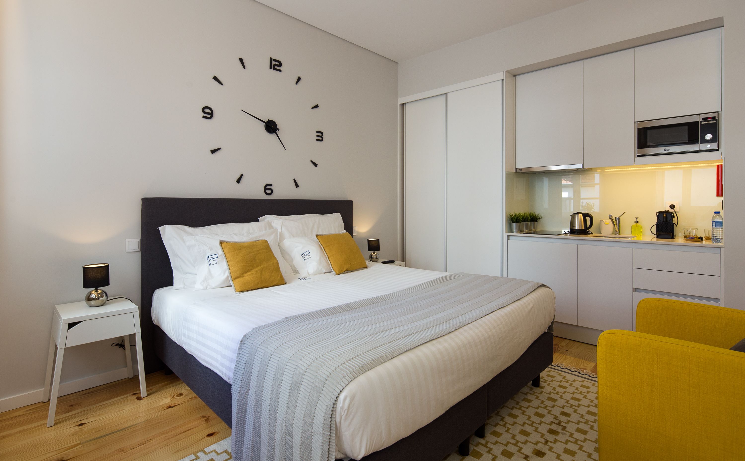 A modern bedroom with a large bed, bedside tables, a clock on the wall, and a kitchenette with a microwave.